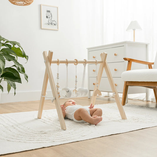 Baby lying on white rug under wooden play gym with hanging plush toys in bright nursery. Come see us at Awaken, Franklin, Tennessee