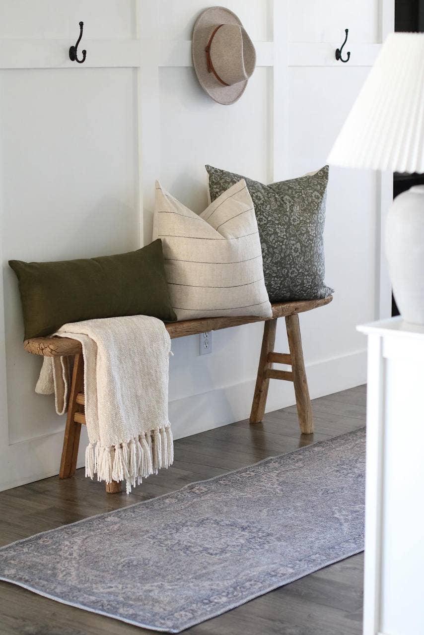 Rustic wooden bench with assorted decorative pillows and beige throw in a minimalist white entryway. Come see us at Awaken, Franklin, Tennessee