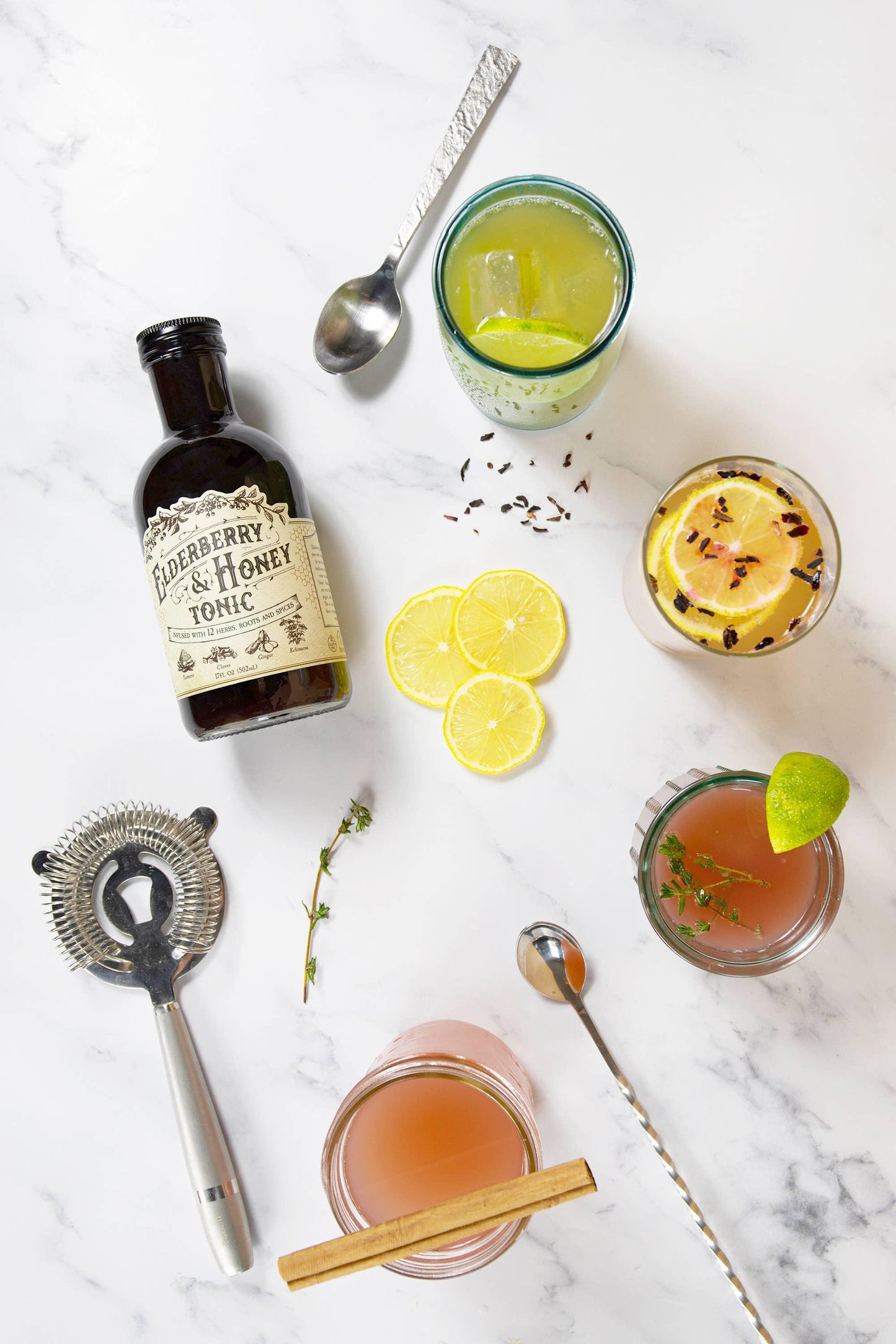 Elderberry & honey tonic bottle with herbal cocktails, citrus slices, bar spoon, and strainer on marble. Come see us at Awaken, Franklin, Tennessee