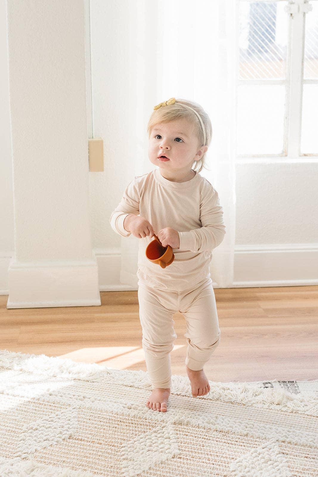 Baby in beige pajamas holds toy cup by window, Come see us at Awaken, Franklin, Tennessee