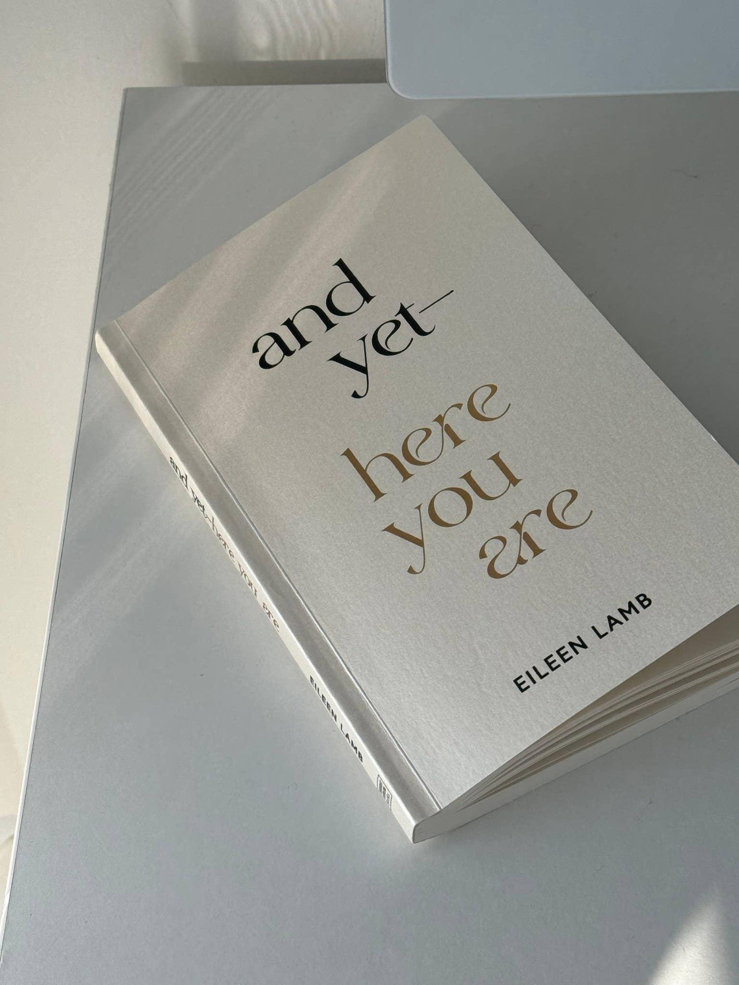 Paperback book titled 'and yet—here you are' by Eileen Lamb on a reflective surface. Come see us at Awaken, Franklin, Tennessee