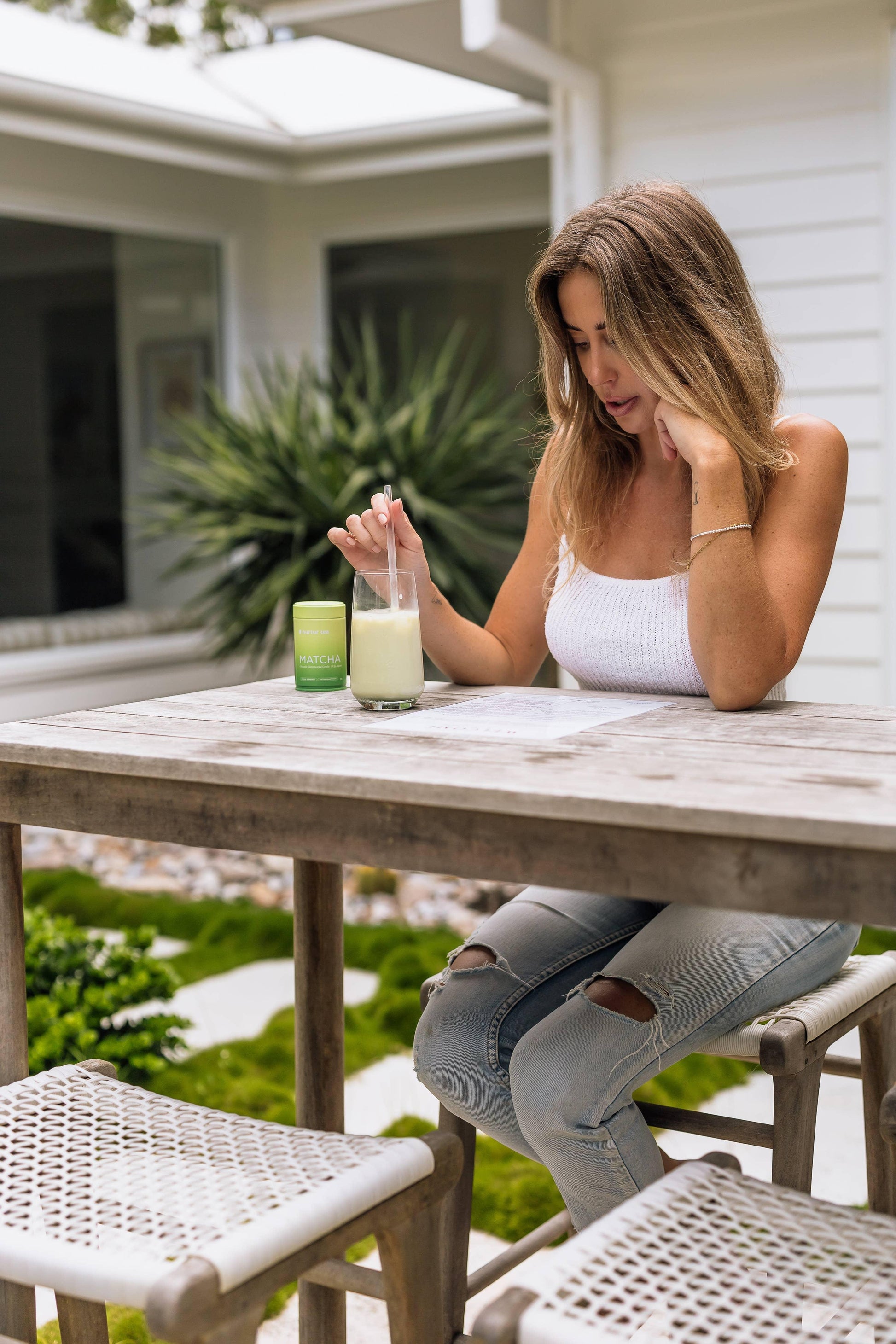 Woman in white tank top and ripped jeans sitting at outdoor wooden table with matcha drink and container. Come see us at Awaken, Franklin, Tennessee