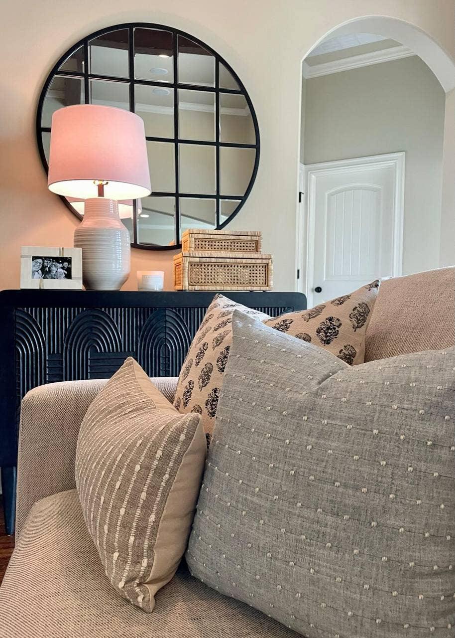 Cozy living room with patterned pillows on beige sofa and round windowpane mirror above black console table. Come see us at Awaken, Franklin, Tennessee