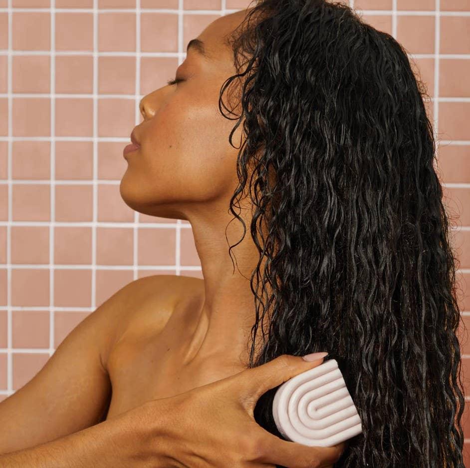 Woman brushing long wet curly hair with pink detangling brush against pink tiled background. Come see us at Awaken, Franklin, Tennessee