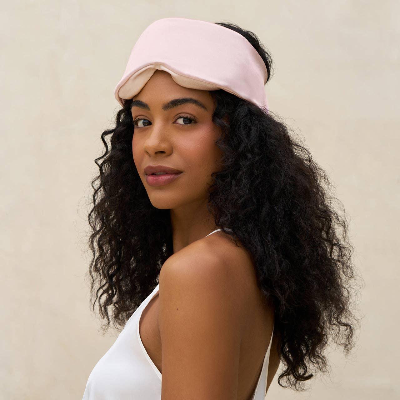 Woman with curly hair wearing pink silk sleep mask and white camisole against neutral background. Come see us at Awaken, Franklin, Tennessee