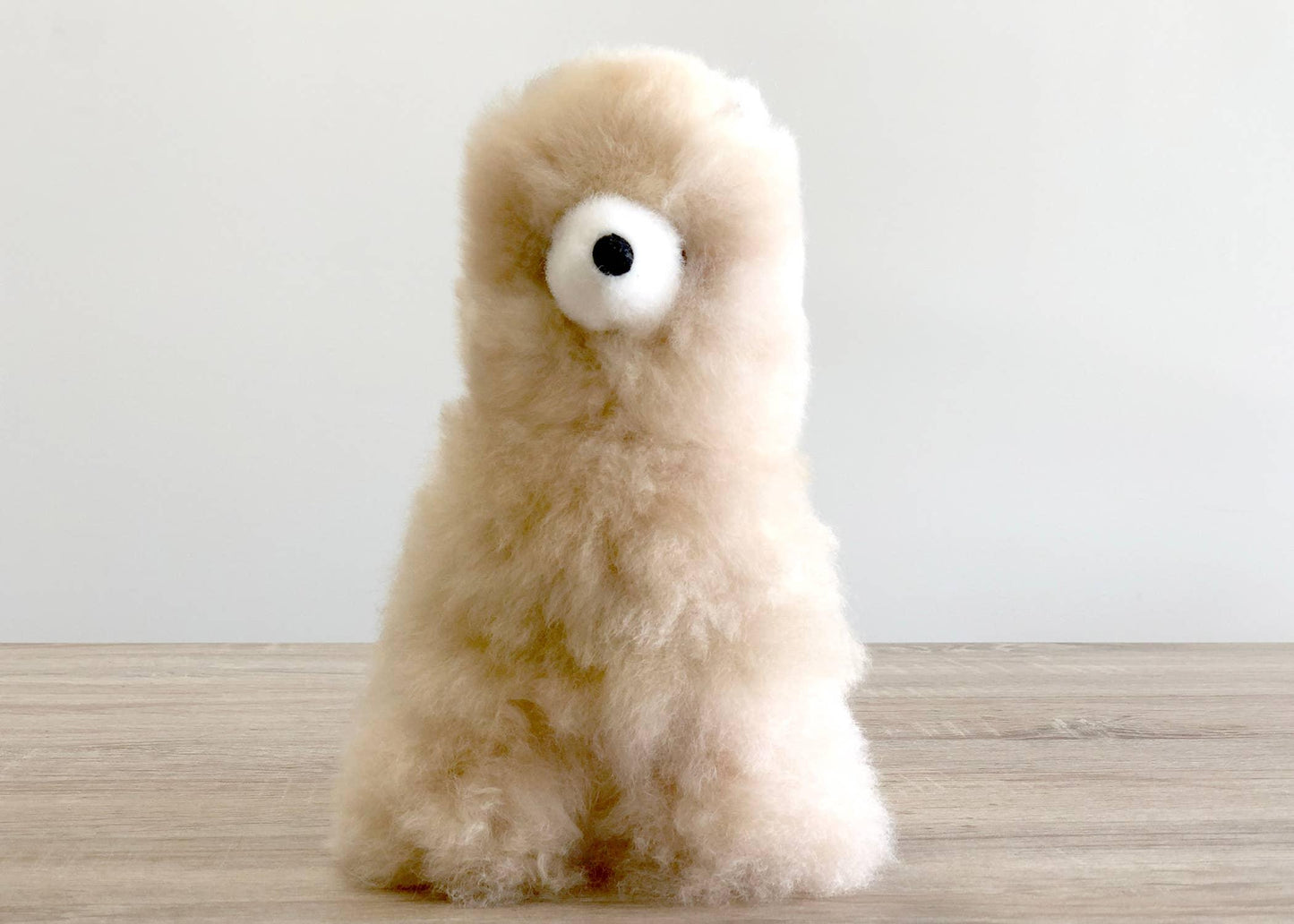 Fluffy beige alpaca plush toy on wooden surface with white background. Come see us at Awaken, Franklin, Tennessee