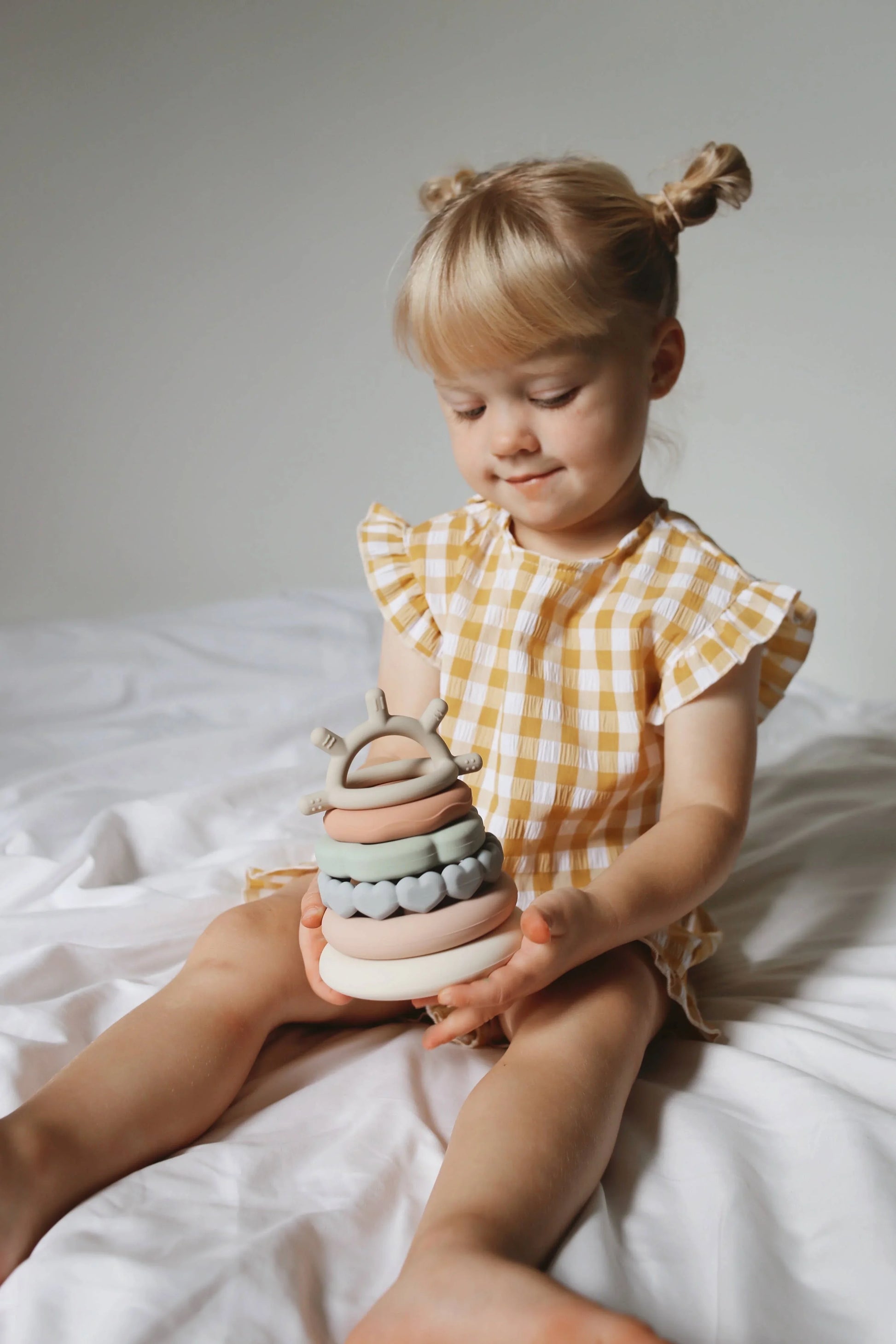 Toddler girl in yellow check dress playing with pastel silicone ring toy on bed. Come see us at Awaken, Franklin, Tennessee