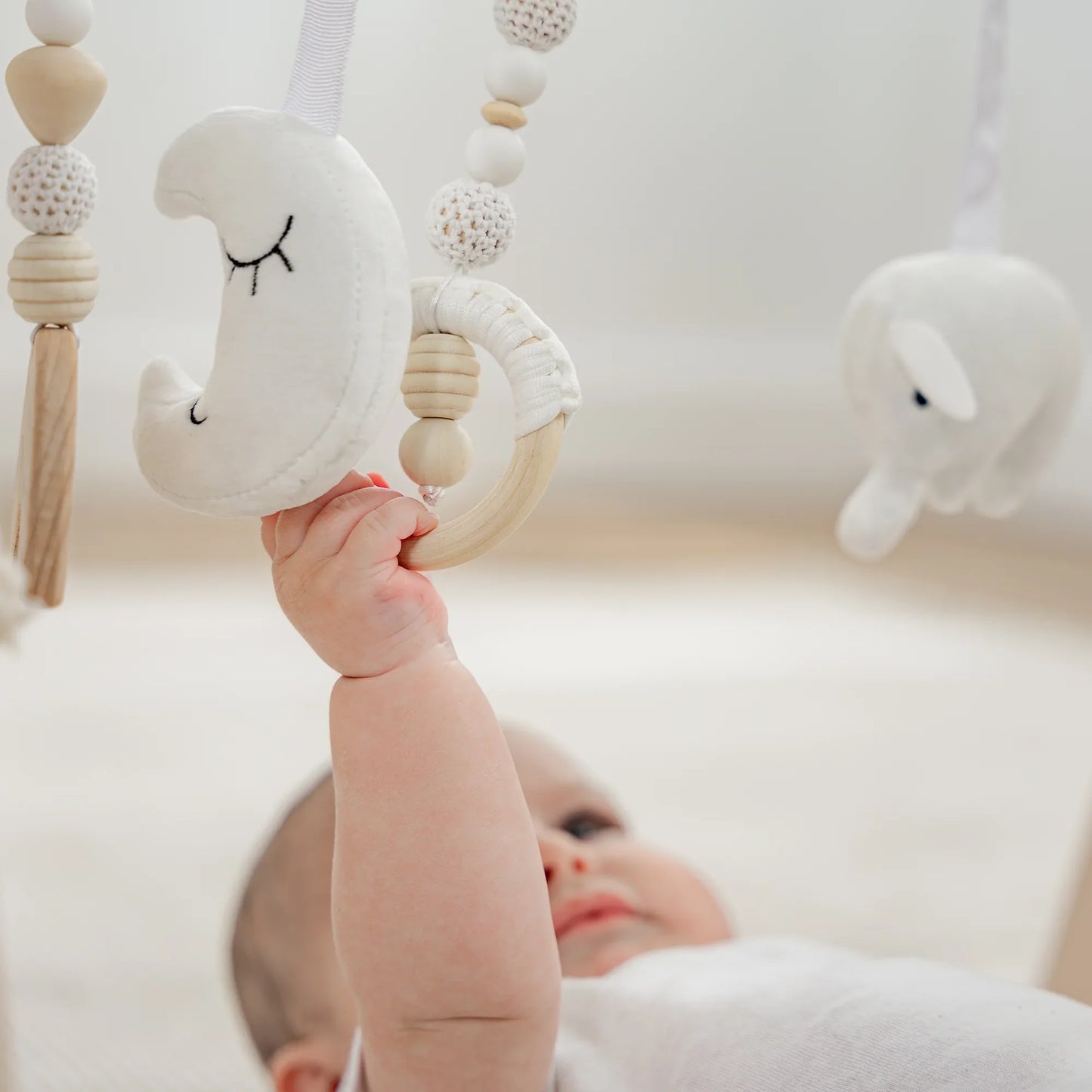Baby lying down grabbing white moon-shaped soft toy on wooden baby gym. Come see us at Awaken, Franklin, Tennessee