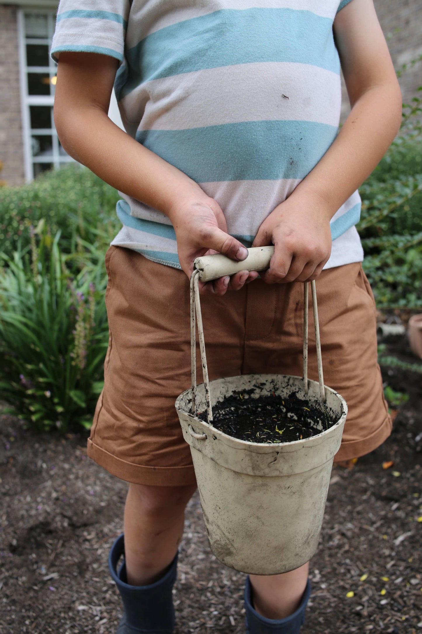 Child holding a dirty white bucket with soil outdoors, wearing striped shirt and brown shorts. Come see us at Awaken, Franklin, Tennessee
