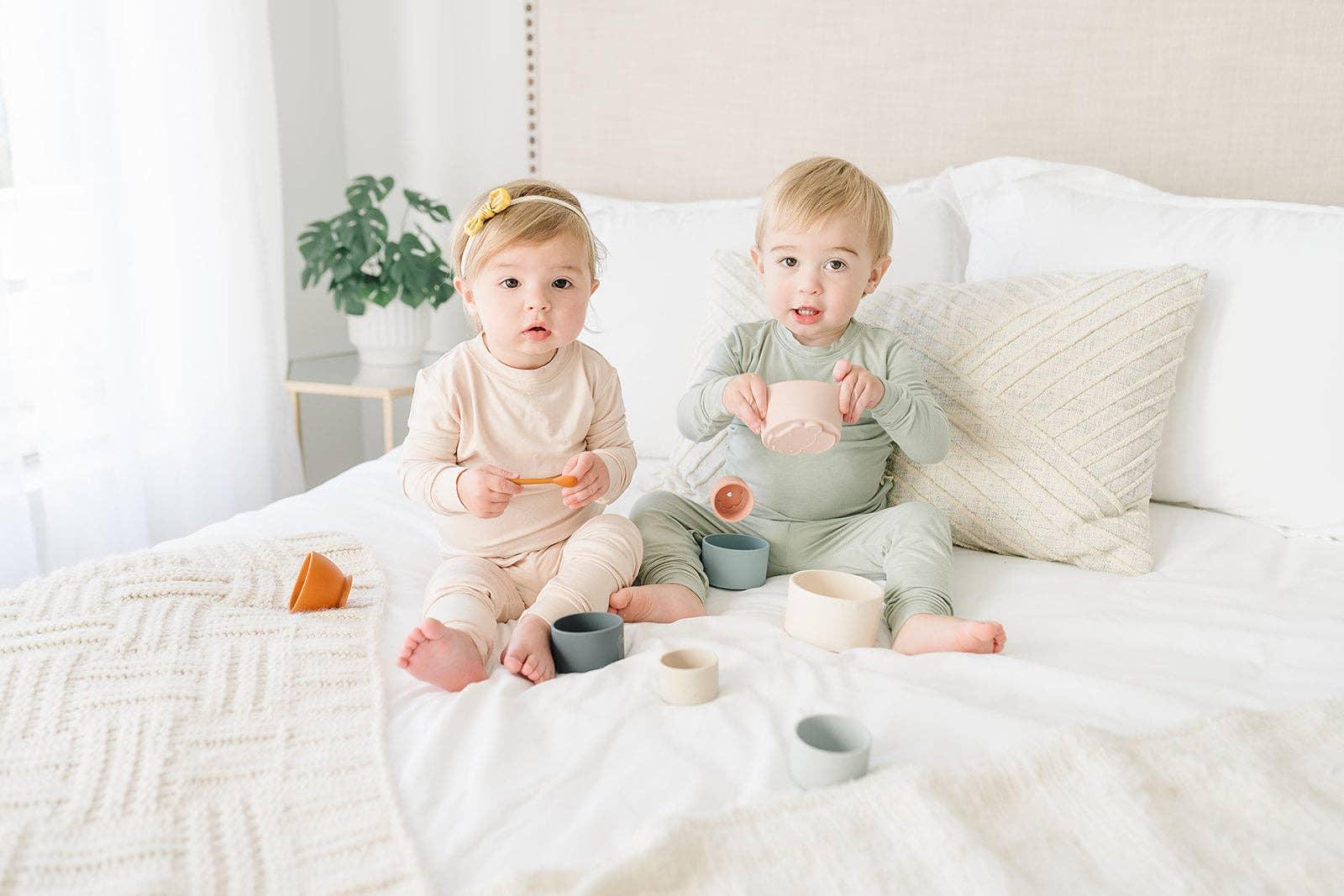 Two toddlers in neutral pajamas sitting on a white bed with soft toys and pillows. Come see us at Awaken, Franklin, Tennessee