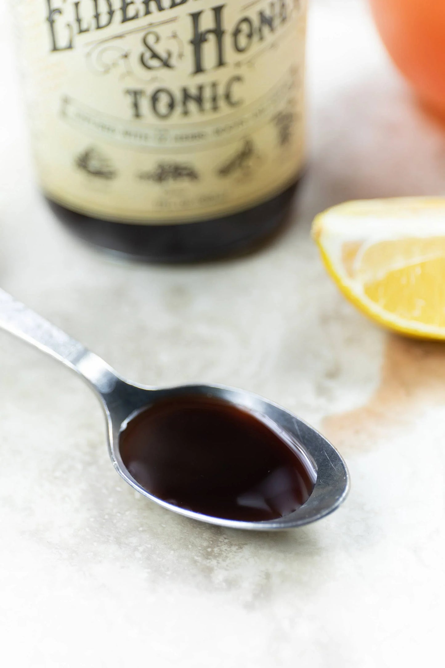 Spoon with dark elderberry and honey tonic syrup, bottle label blur and lemon wedge nearby. Come see us at Awaken, Franklin, Tennessee