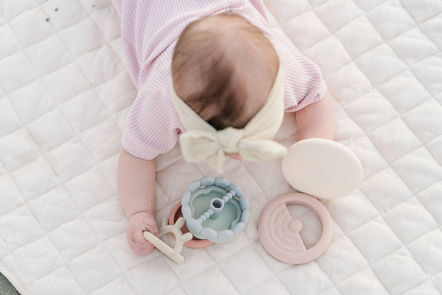 Baby in pink with headband playing with pastel silicone toys on quilted mat. Come see us at Awaken, Franklin, Tennessee