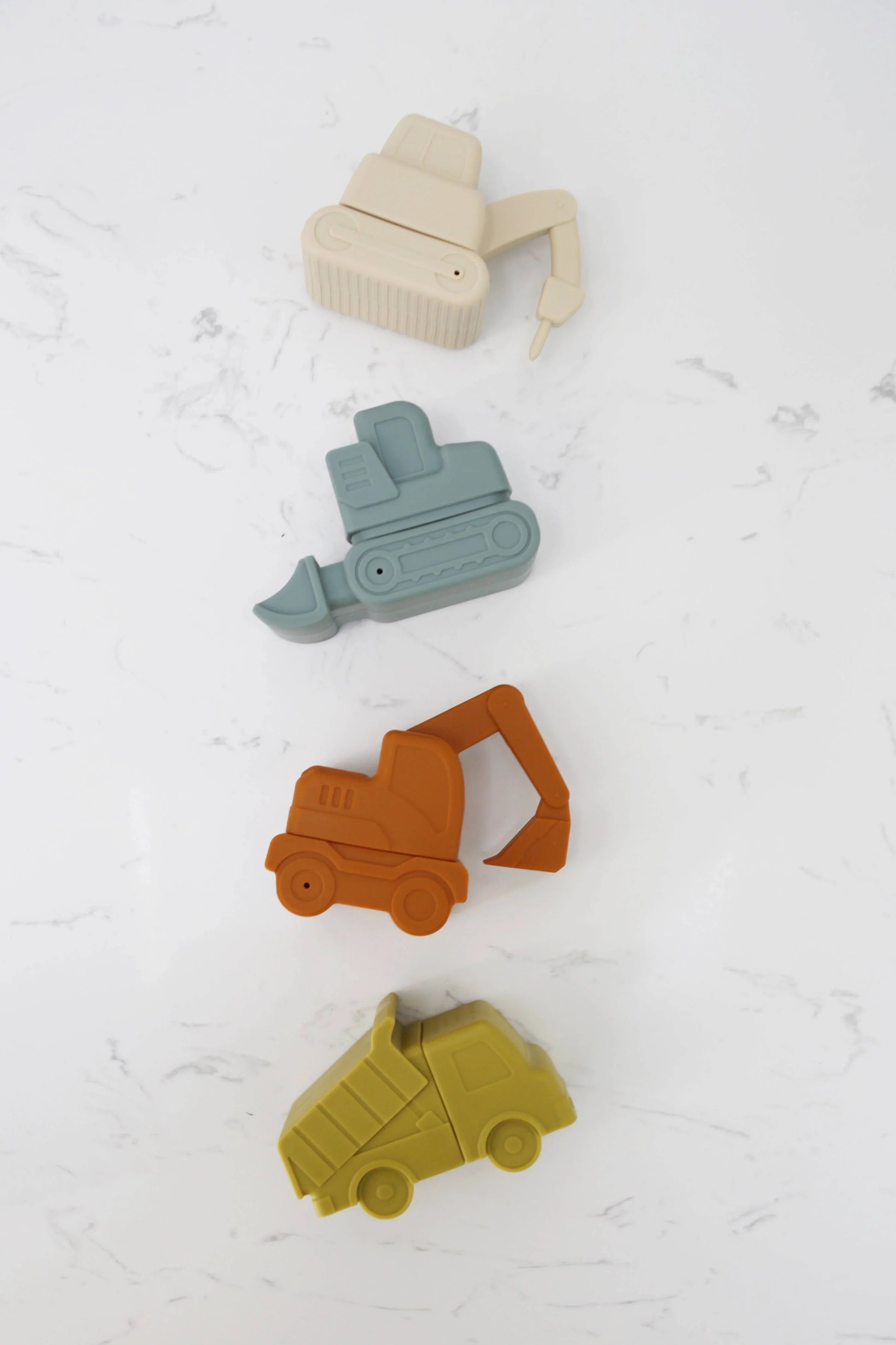 Silicone toy construction vehicles in beige, blue, orange, and yellow on white marble background, Come see us at Awaken, Franklin, Tennessee