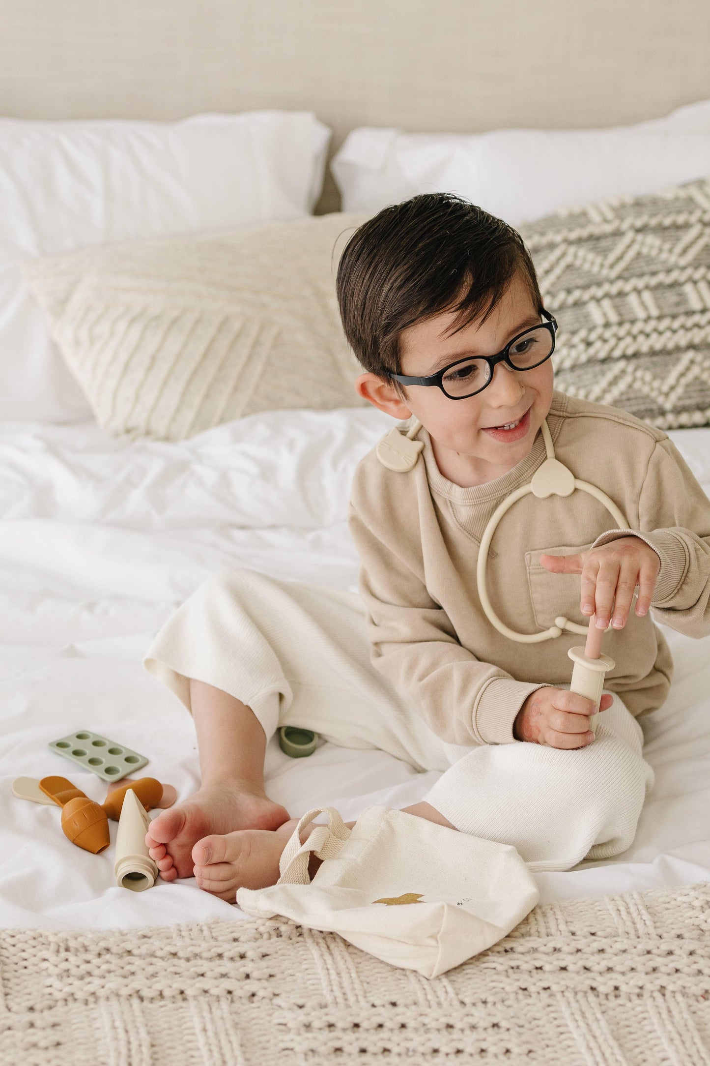 Smiling young boy with glasses playing with beige and brown toy medical set on white bed Come see us at Awaken, Franklin, Tennessee