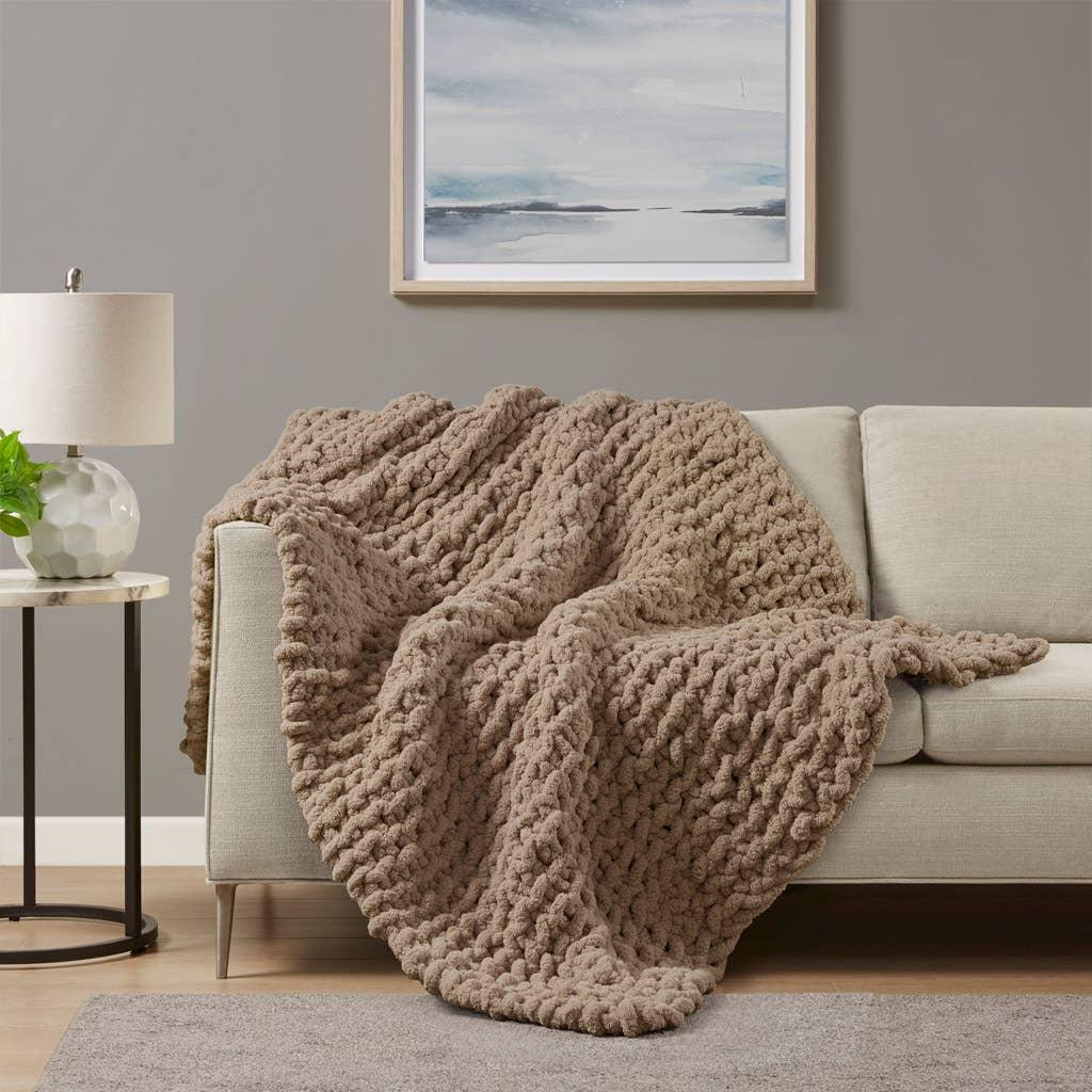 Chunky knit beige throw blanket draped over light sofa in cozy living room with lamp and abstract art. Come see us at Awaken, Franklin, Tennessee