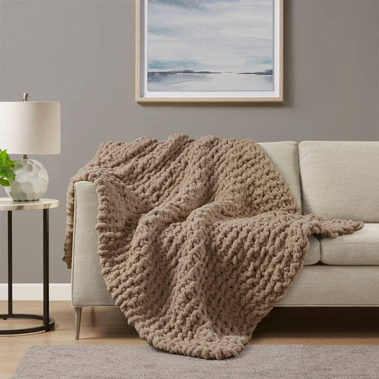 Chunky knit beige throw blanket draped over light sofa in cozy living room with lamp and abstract art. Come see us at Awaken, Franklin, Tennessee