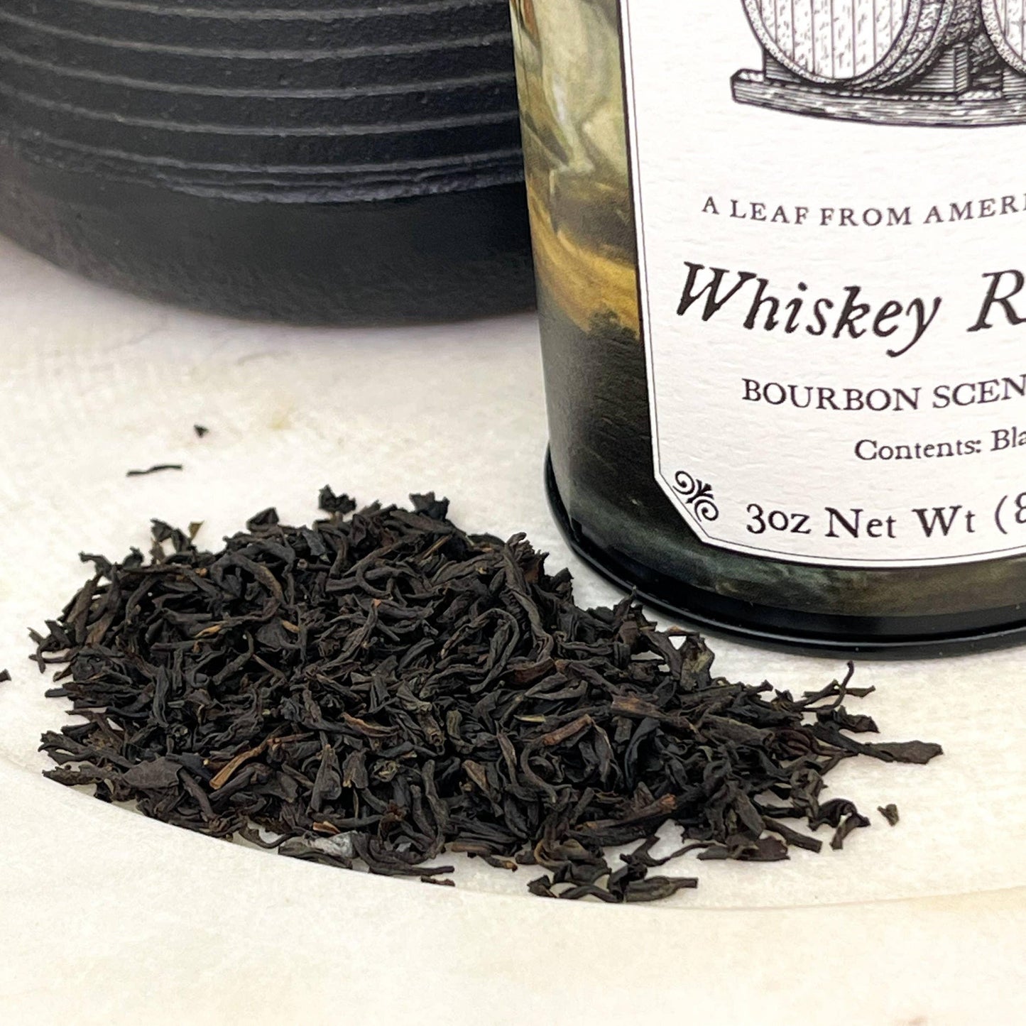 Loose black tea leaves next to a whiskey rum bourbon scented candle jar on white surface. Come see us at Awaken, Franklin, Tennessee