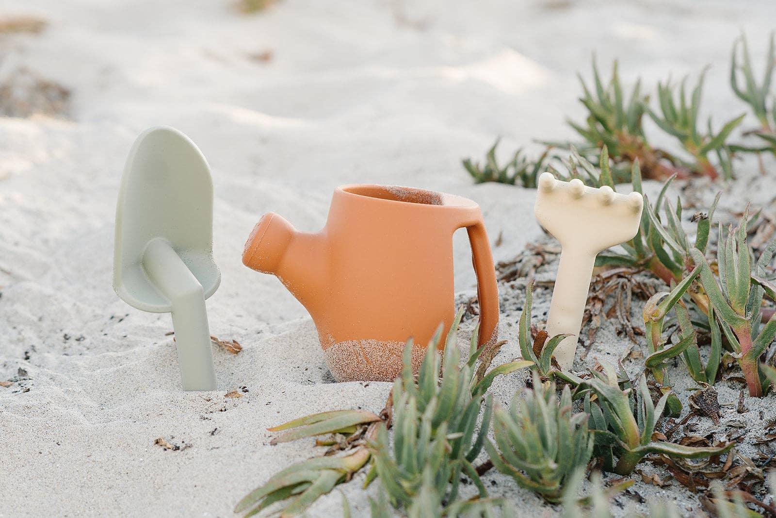 Child's gardening tools including a green trowel, orange watering can, and white rake in sandy soil with green plants. Come see us at Awaken, Franklin, Tennessee