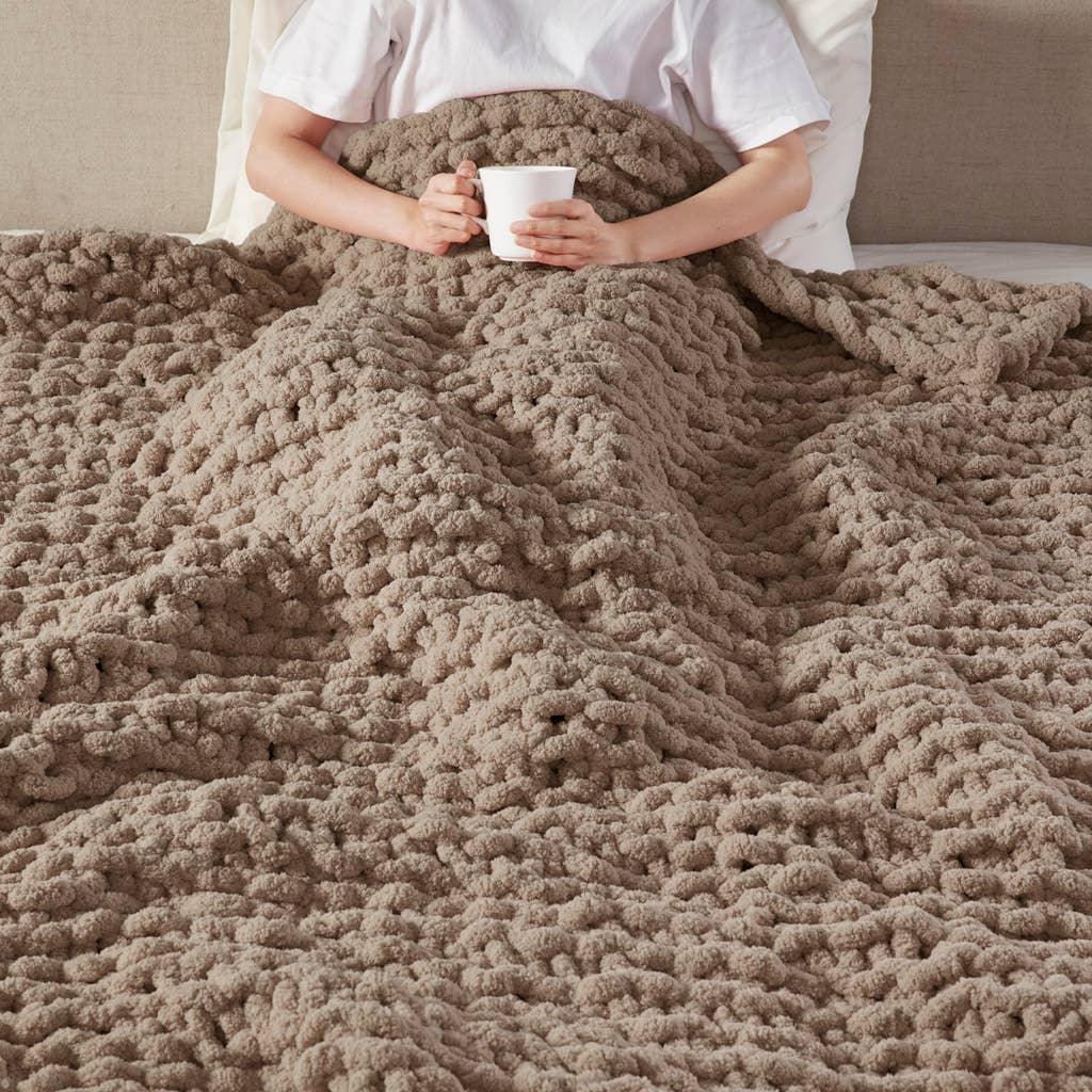 Person sitting in bed wrapped in chunky knit beige blanket holding white mug, Come see us at Awaken, Franklin, Tennessee