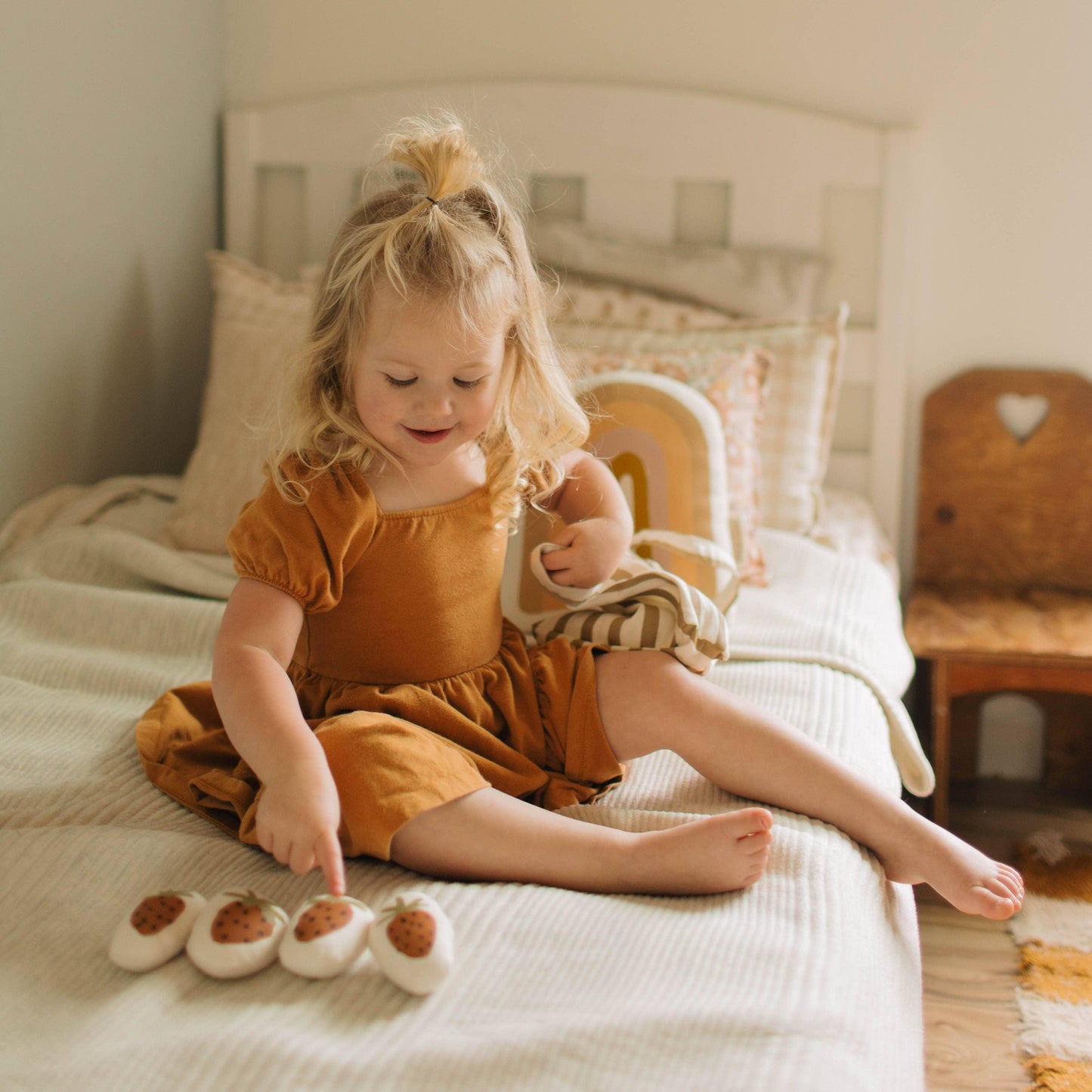 Smiling toddler girl in mustard dress on bed with toys, cozy decor. Come see us at Awaken, Franklin, Tennessee