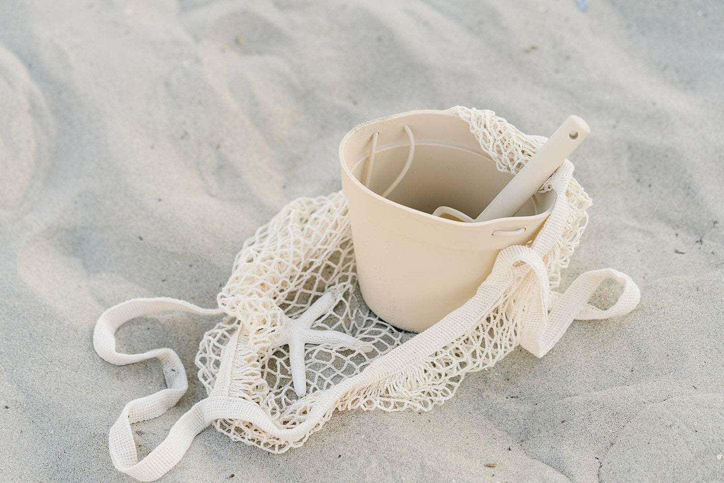 Beach sand with a beige bucket, shovel, starfish, and a mesh bag. Come see us at Awaken, Franklin, Tennessee