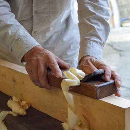 Hands planing wood with a hand plane creating shavings, woodworker wearing white shirt. Come see us at Awaken, Franklin, Tennessee