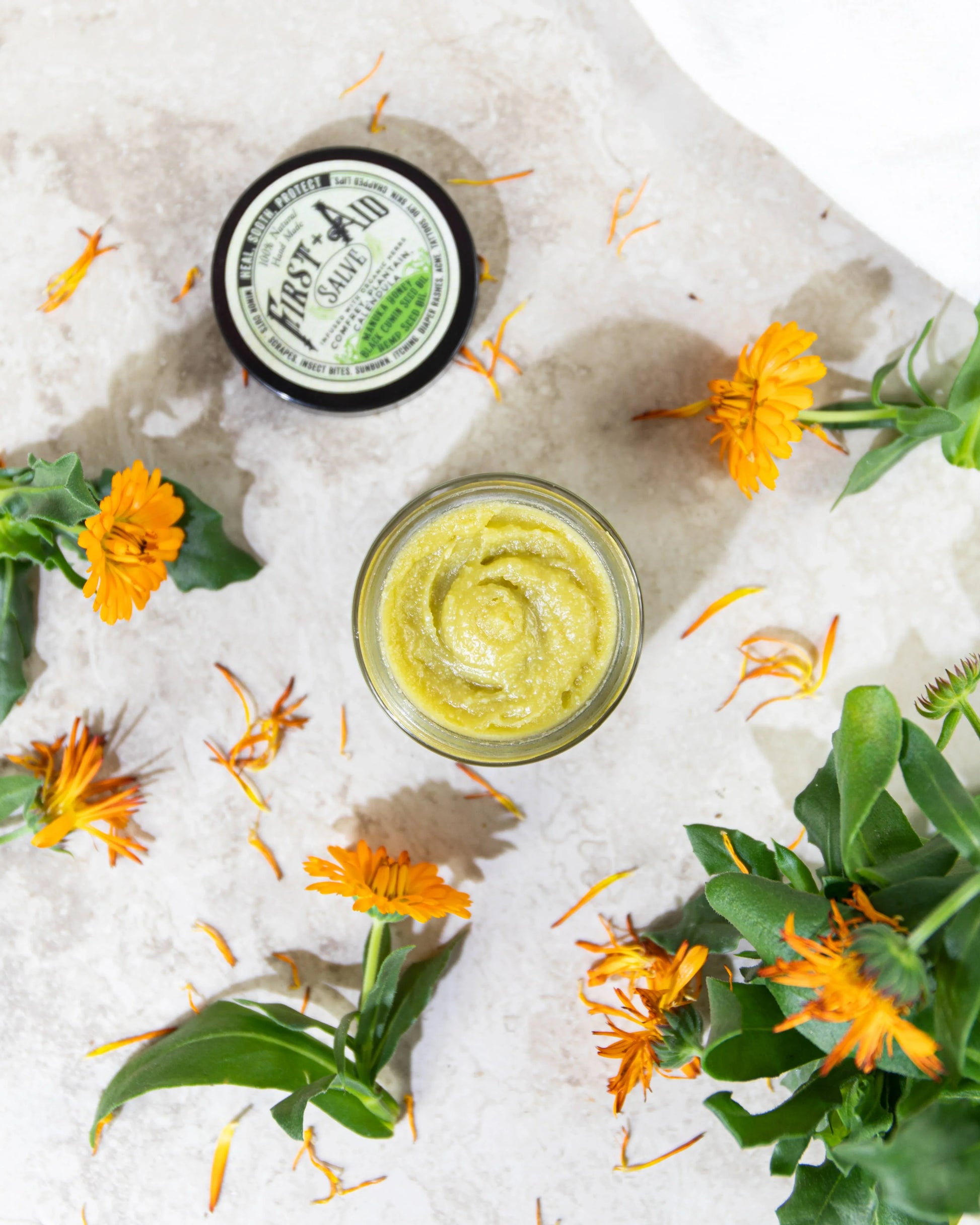 Open jar of First Aid Salve with calendula flowers on marble surface, natural skincare. Come see us at Awaken, Franklin, Tennessee