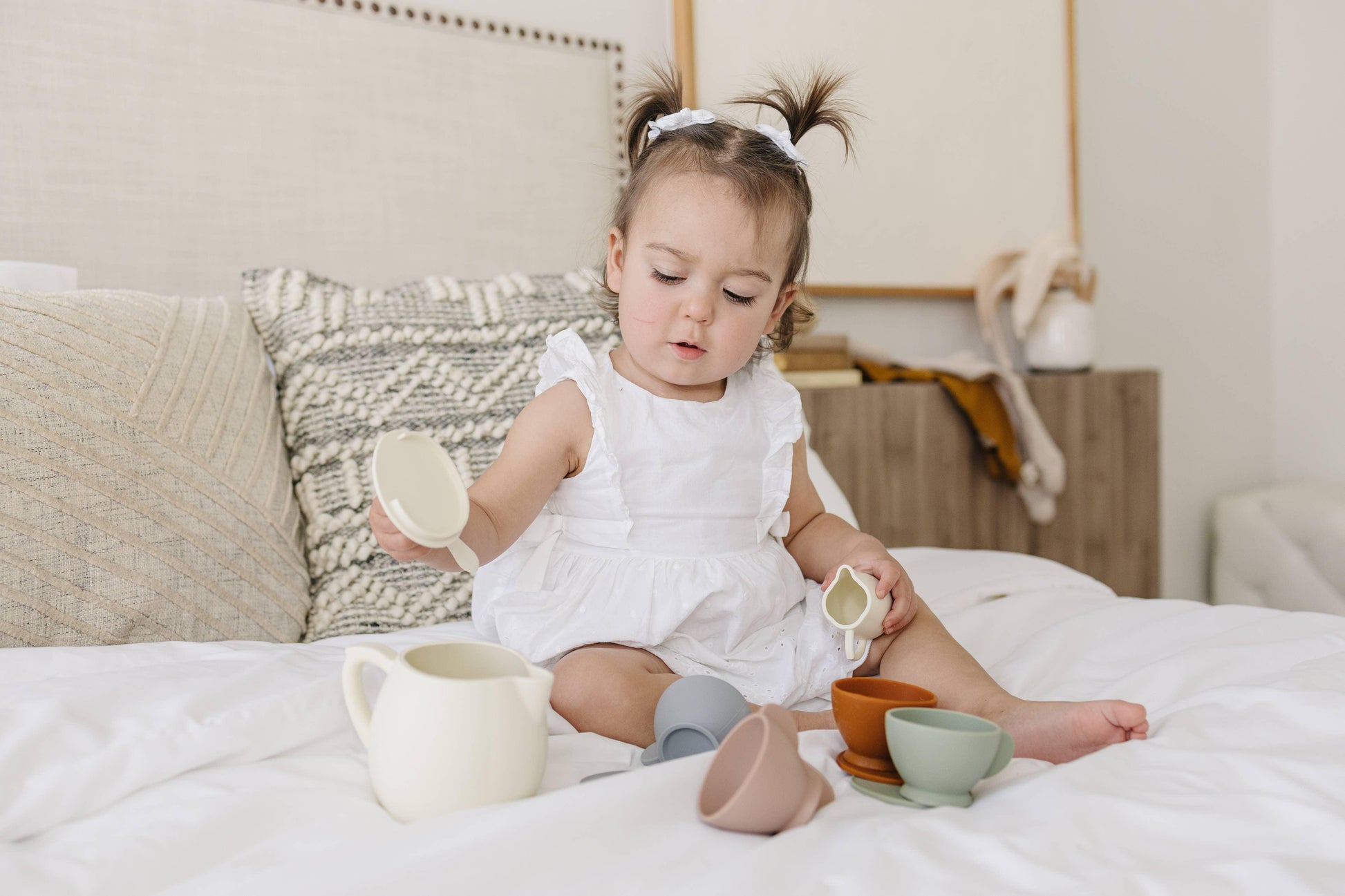 Toddler girl in white dress plays with toy tea set on bed, Come see us at Awaken, Franklin, Tennessee