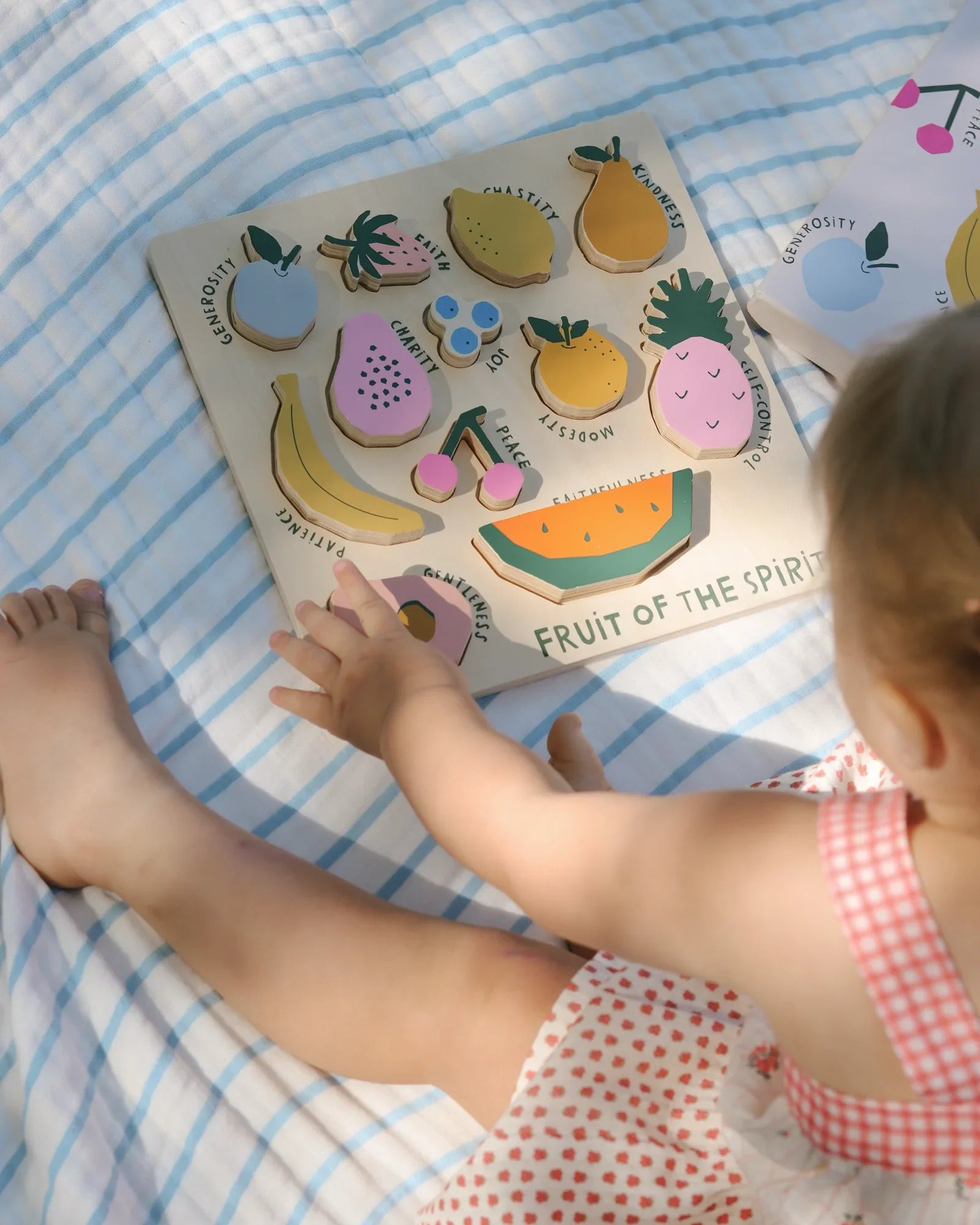 Toddler playing with wooden Fruit of the Spirit puzzle on blue-striped blanket. Come see us at Awaken, Franklin, Tennessee