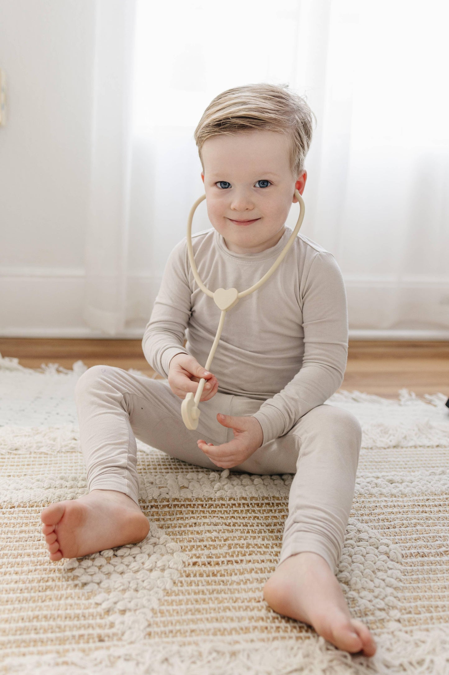 Young boy sitting on textured rug playing with beige heart-shaped stethoscope, bright room Come see us at Awaken, Franklin, Tennessee
