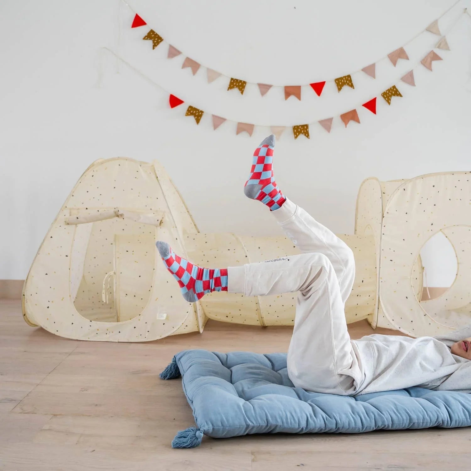 Child lying on blue floor cushion wearing red and blue checkered socks in playroom with beige fabric play tents and banners. Come see us at Awaken, Franklin, Tennessee