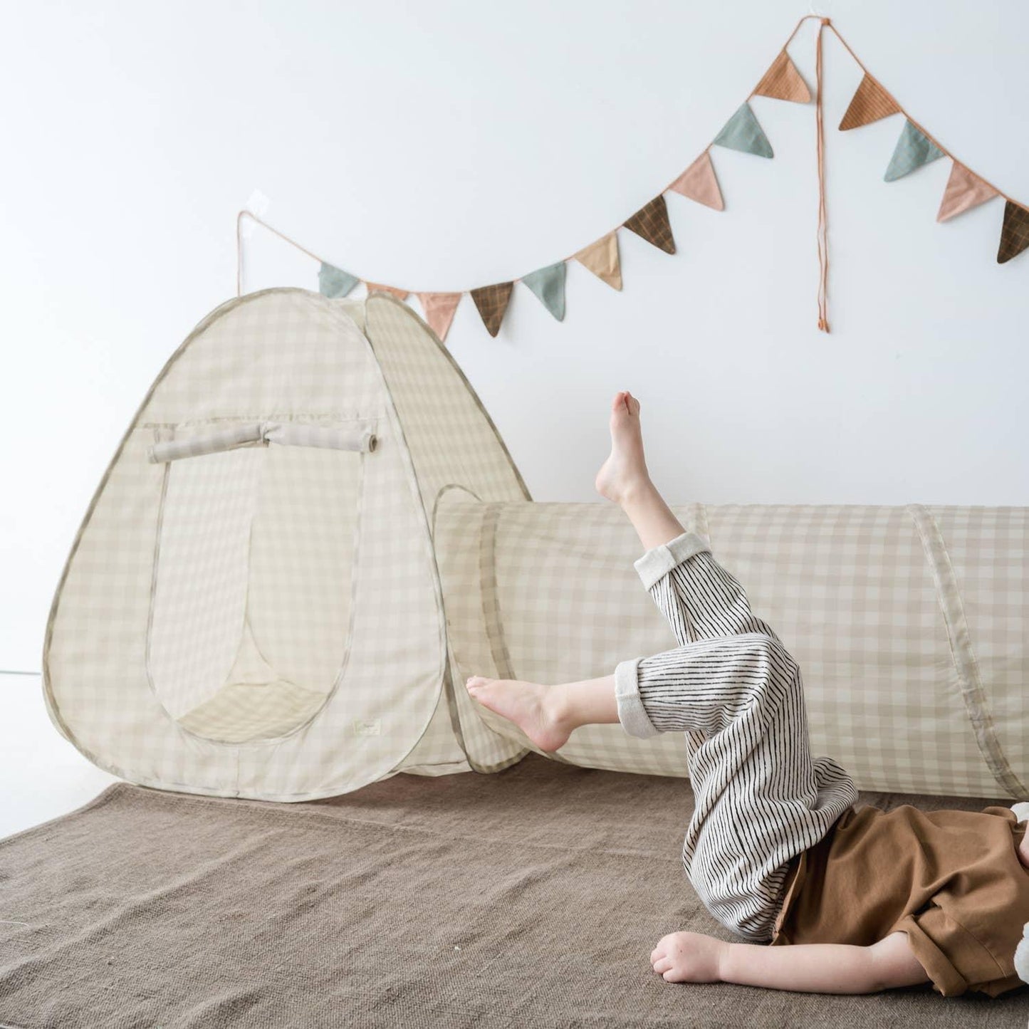 Child playing in beige checkered indoor play tent and tunnel on brown rug with bunting flags. Come see us at Awaken, Franklin, Tennessee