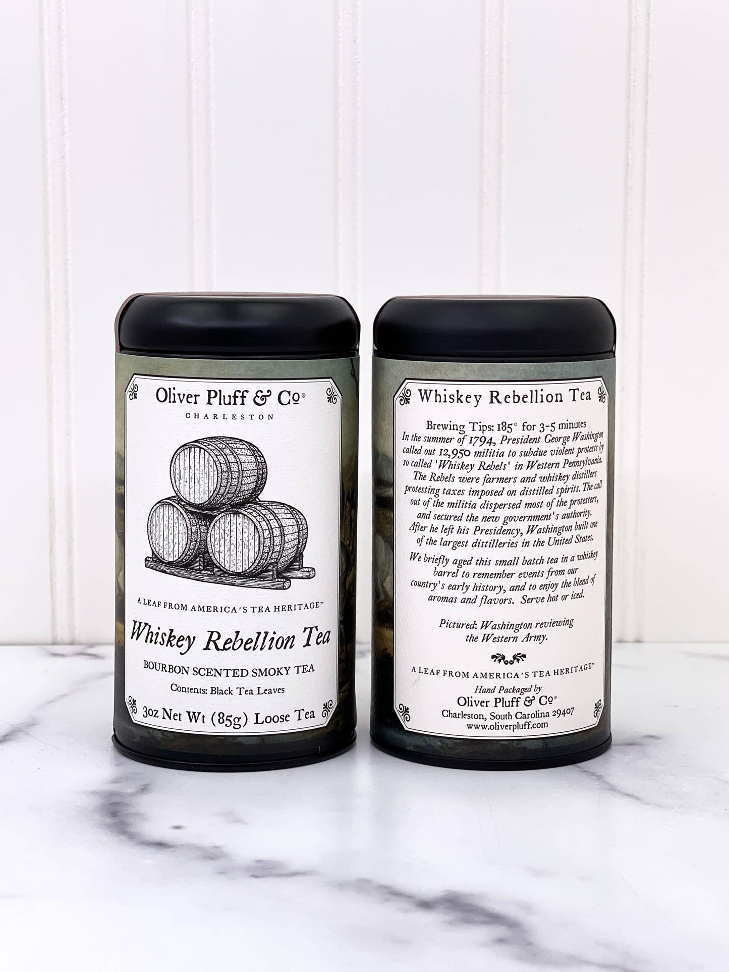 Two olive green tins of Whiskey Rebellion Tea by Oliver Pluff & Co on marble surface, with black lids. Come see us at Awaken, Franklin, Tennessee