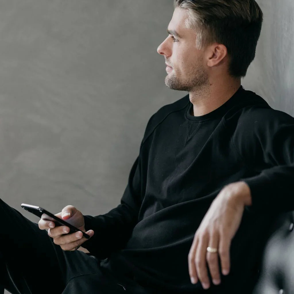 Young man in black sweatshirt holding phone, sitting against gray wall, relaxed pose. Come see us at Awaken, Franklin, Tennessee