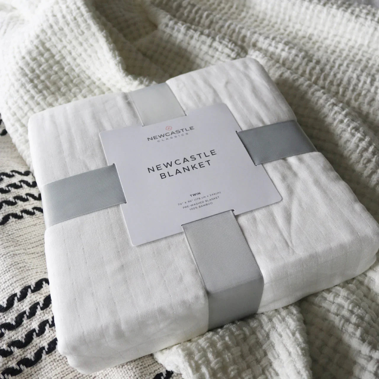 Newcastle Classics white twin bamboo blanket wrapped in gray ribbon on textured fabric Come see us at Awaken, Franklin, Tennessee