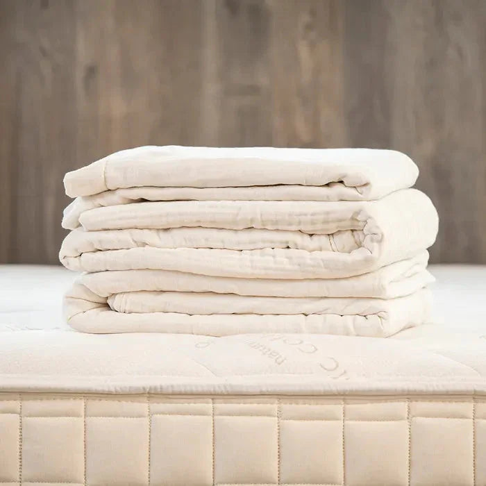 Folded beige organic cotton mattress topper on a quilted mattress with wooden background. Come see us at Awaken, Franklin, Tennessee