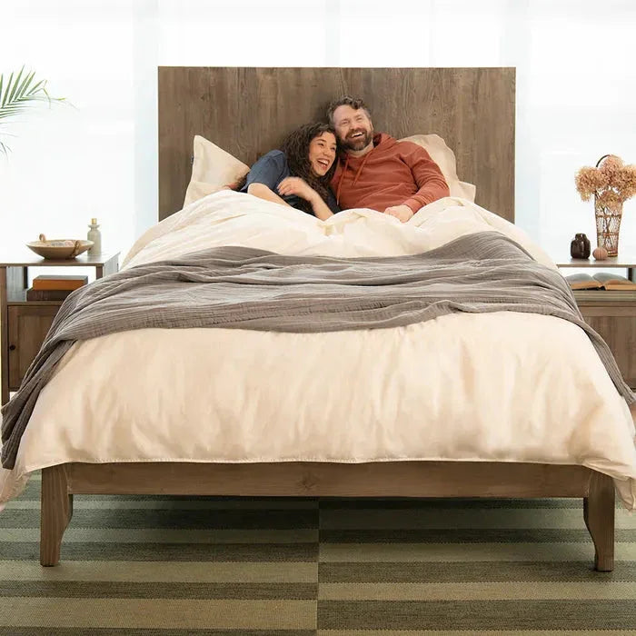 Happy couple relaxing in a cozy wooden bed with beige bedding and gray throw in bright bedroom. Come see us at Awaken, Franklin, Tennessee