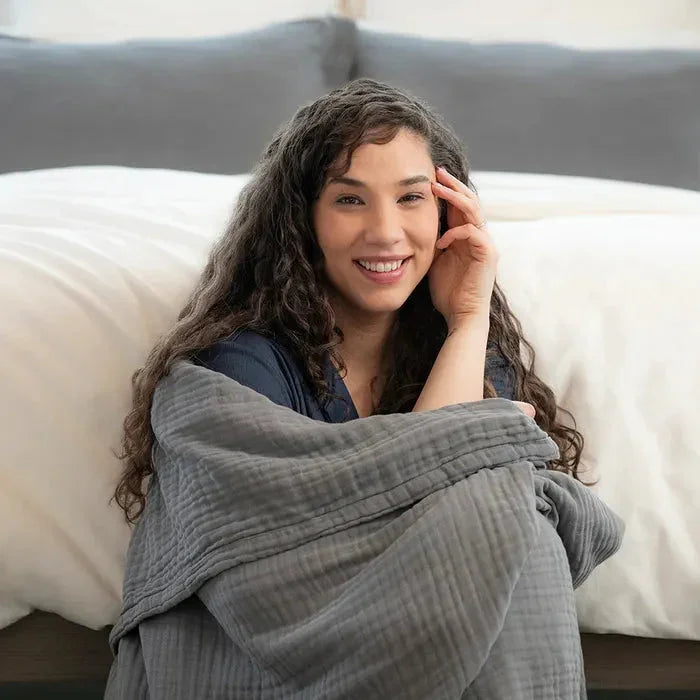 Smiling woman wrapped in cozy gray blanket sitting on bed pillows. Come see us at Awaken, Franklin, Tennessee