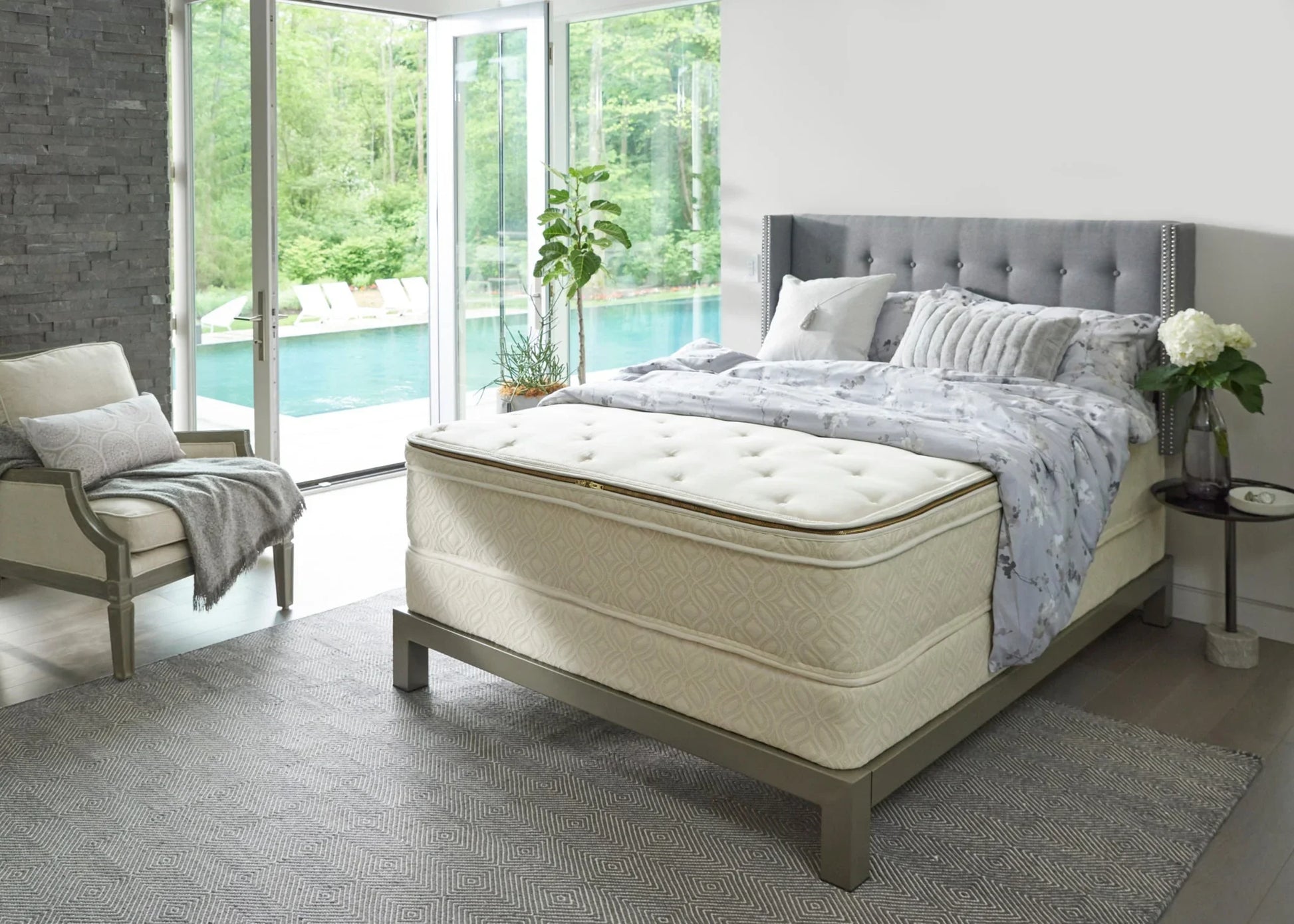 Modern bedroom with tufted gray bed, floral bedding, beige armchair, overlooking pool and greenery. Come see us at Awaken, Franklin, Tennessee