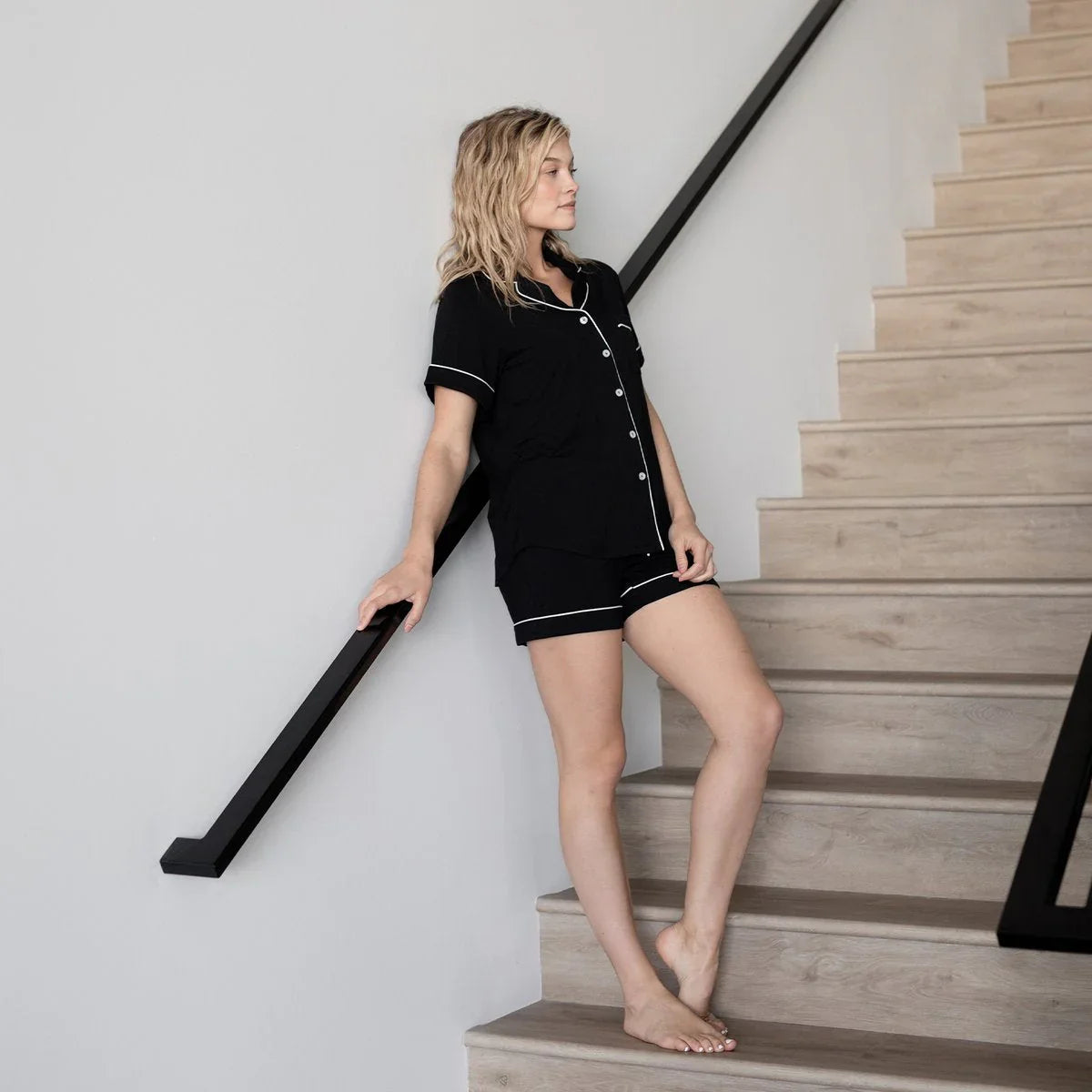 Woman in black pajama set with white piping standing barefoot on wooden stairs, minimal white wall background. Come see us at Awaken, Franklin, Tennessee