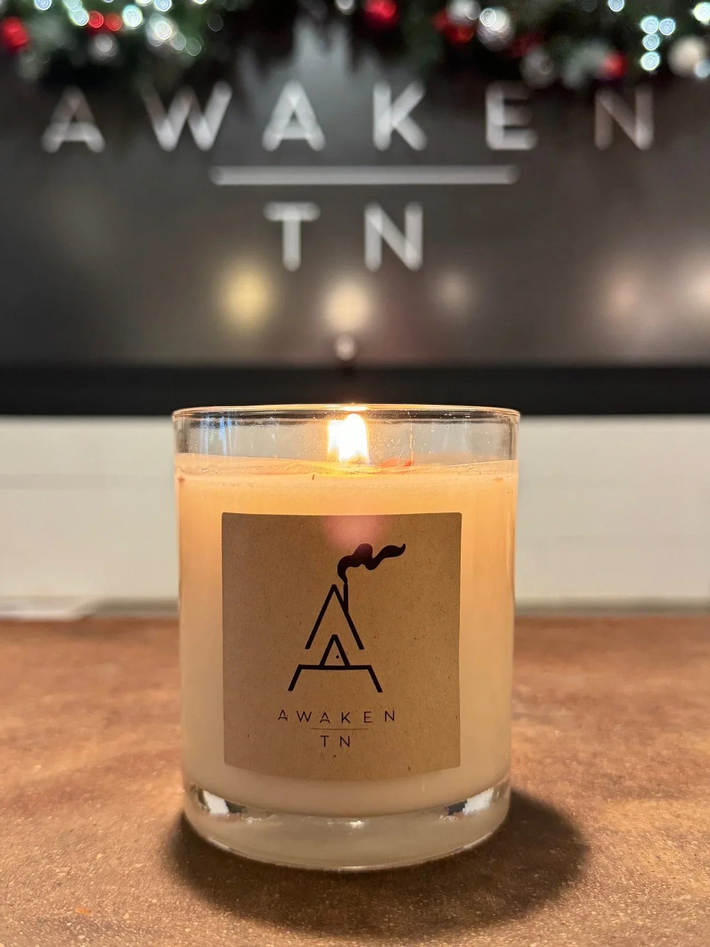 Lit vintage-style candle with Awaken TN logo on label and blurred Awaken sign above, Come see us at Awaken, Franklin, Tennessee