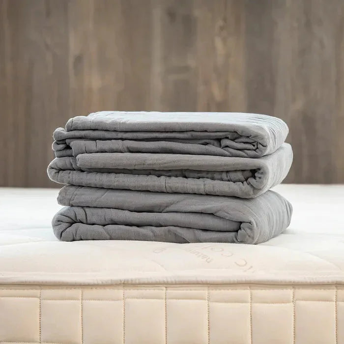 Stack of soft gray weighted blankets on a mattress with wooden wall background. Come see us at Awaken, Franklin, Tennessee