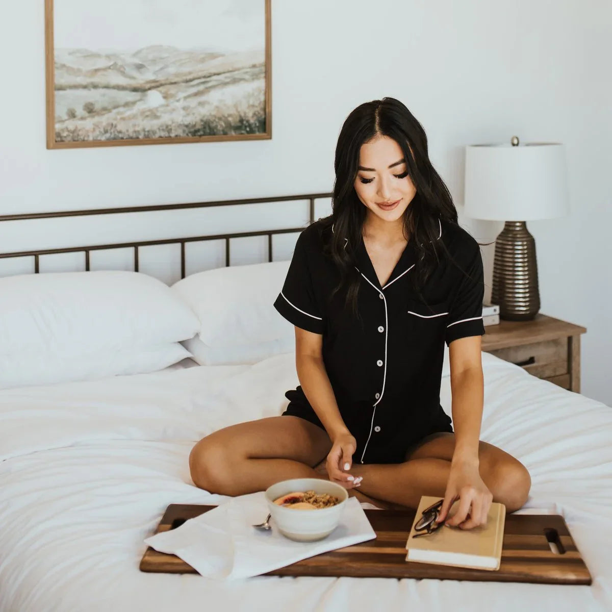 Woman in black pajamas sitting cross-legged on bed with breakfast tray, lamp and painting in room. Come see us at Awaken, Franklin, Tennessee